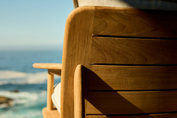 Close‑up of a honey brown teak outdoor lounge chair back with wide horizontal slats and rounded corners facing the coast
