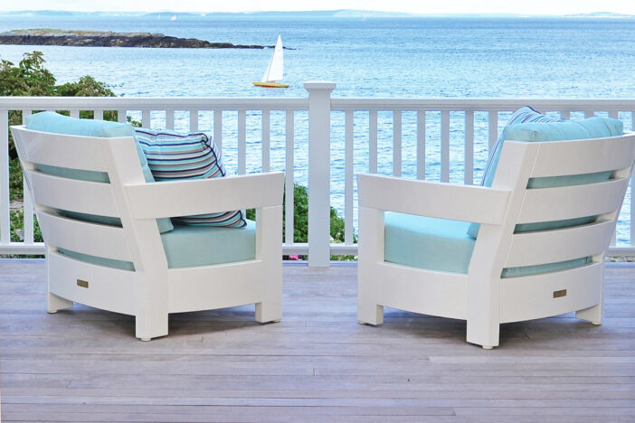 Stout outdoor lounge chairs with thick white slat frames and glossy finish with light teal cushions on a veranda overlooking the coast