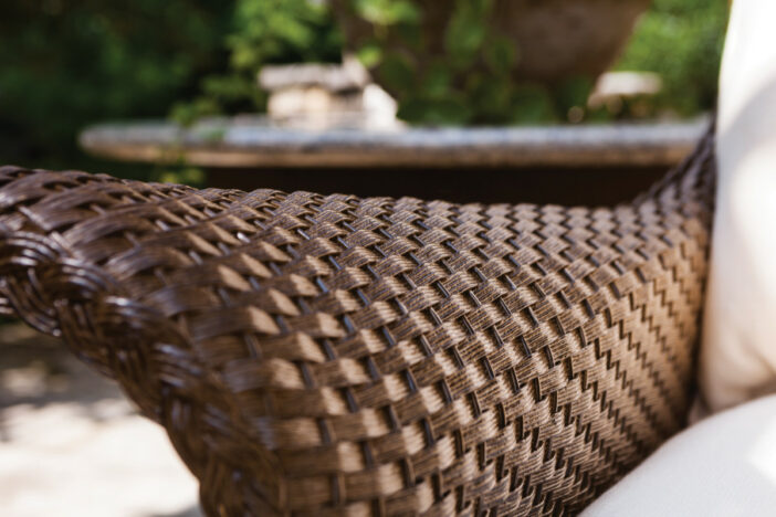 Close‑up of the flared arm of a dark brown outdoor wicker lounge chair with white cushions and trees in the background