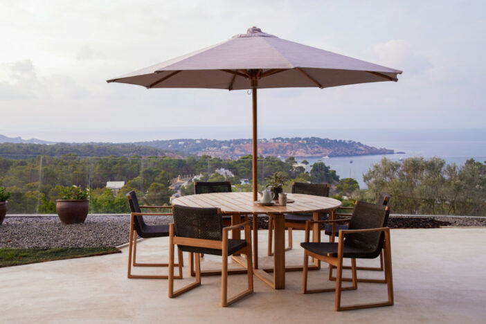 Octagonal center pole umbrella thru a teak dining table surrounded by chairs on a coastal patio