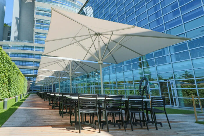 Line of large white center pole umbrellas with square canopies between dining sets on a corporate terrace