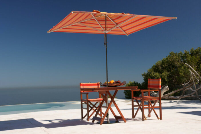 Center pole umbrella with bright orange and red striped square canopy through a teak folding table on a coastal patio