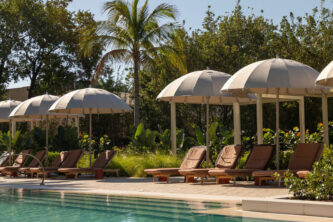 Beige center pole umbrellas with tasseled rounded canopies between brown sun loungers alongside a resort pool