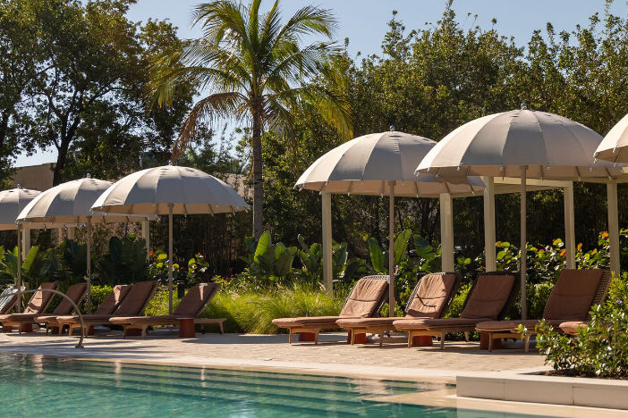 Beige center pole umbrellas with tasseled rounded canopies between brown sun loungers alongside a resort pool
