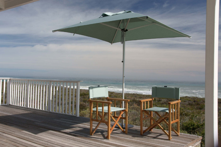 Silver market umbrella with a square light teal canopy between two folding wooden armchairs on a coastal deck